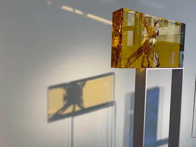 Modern art sculpture with amber resin block casting shadow on gallery wall, showcasing contemporary design and artistic display.